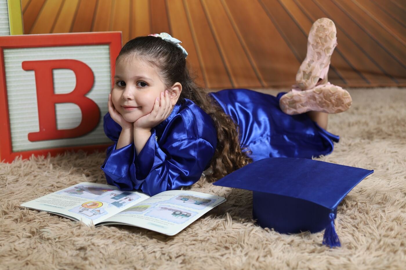 Menina de beca azul deitada no tapete, lendo ao lado do cubo B